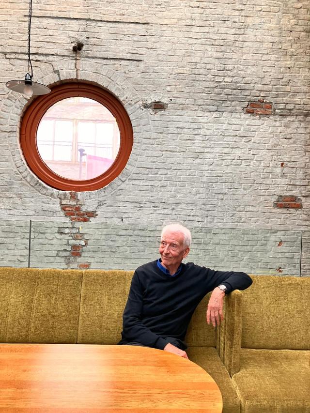 Portret van Jan Vanderhaeghe in een groenen zetel. Hij leunt met zijn linkerarm ontspannen op de rugleuning en kijkt glimlachend naar een punt rechts van de camera.