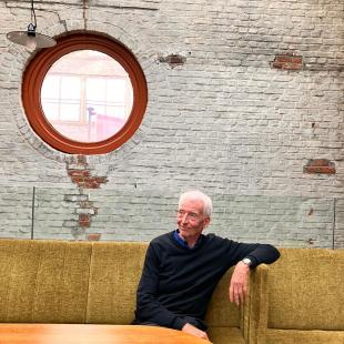 Portret van Jan Vanderhaeghe in een groene zetel. Hij leunt met zijn linkerarm ontspannen op de rugleuning en kijkt glimlachend naar een punt rechts van de camera.