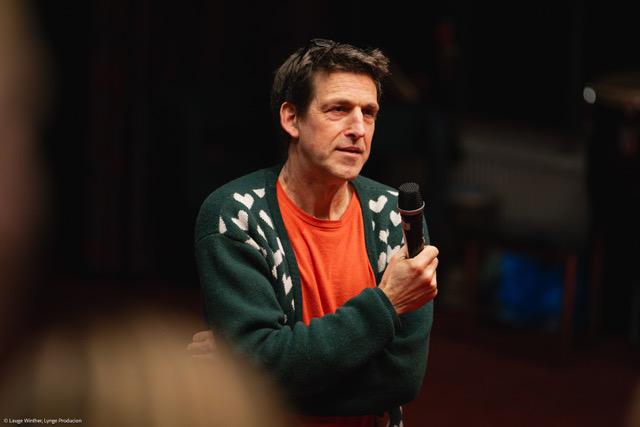 Dries Meddens met een microfoon in zijn hand, tegen een zwarte achtergrond.